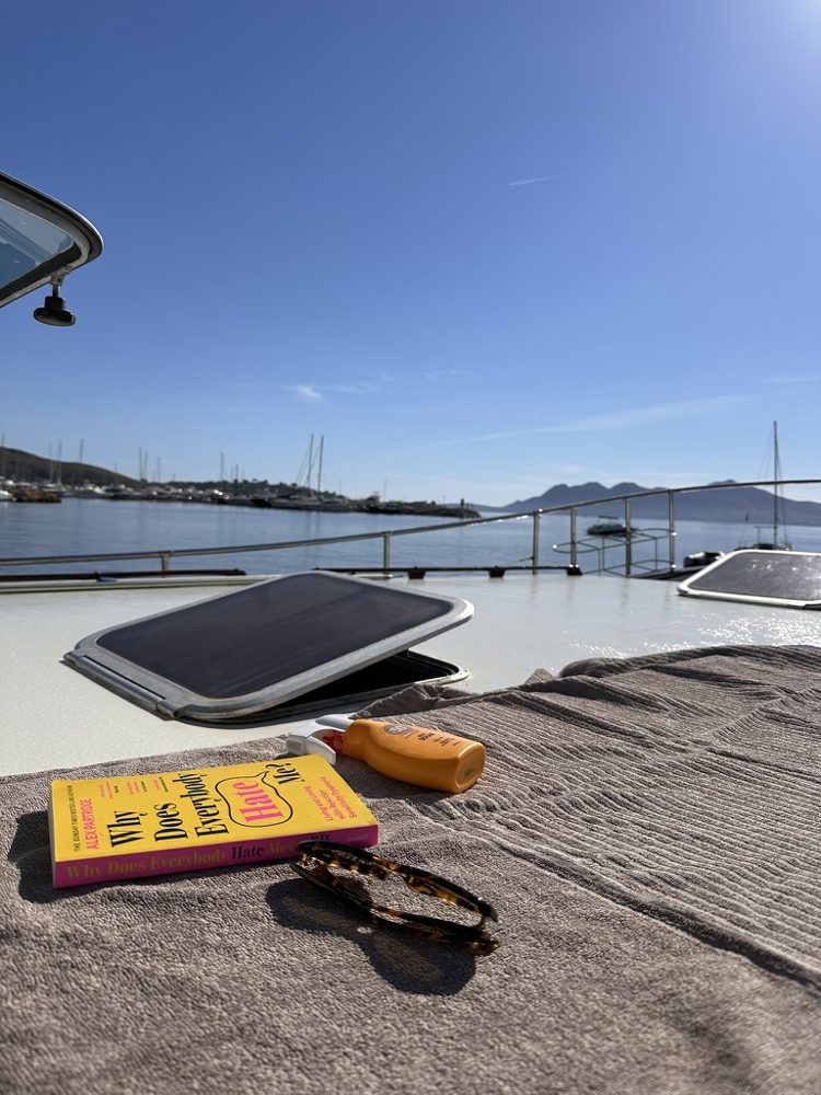 Why Does Everybody Hate Me? by Alex Partridge on a towel on a boat deck in Mallorca, next to sunscreen and sunglasses